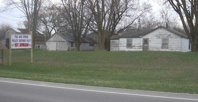 Run-down looking house and gray farm outbuildings with pristine handmade sign in front yard