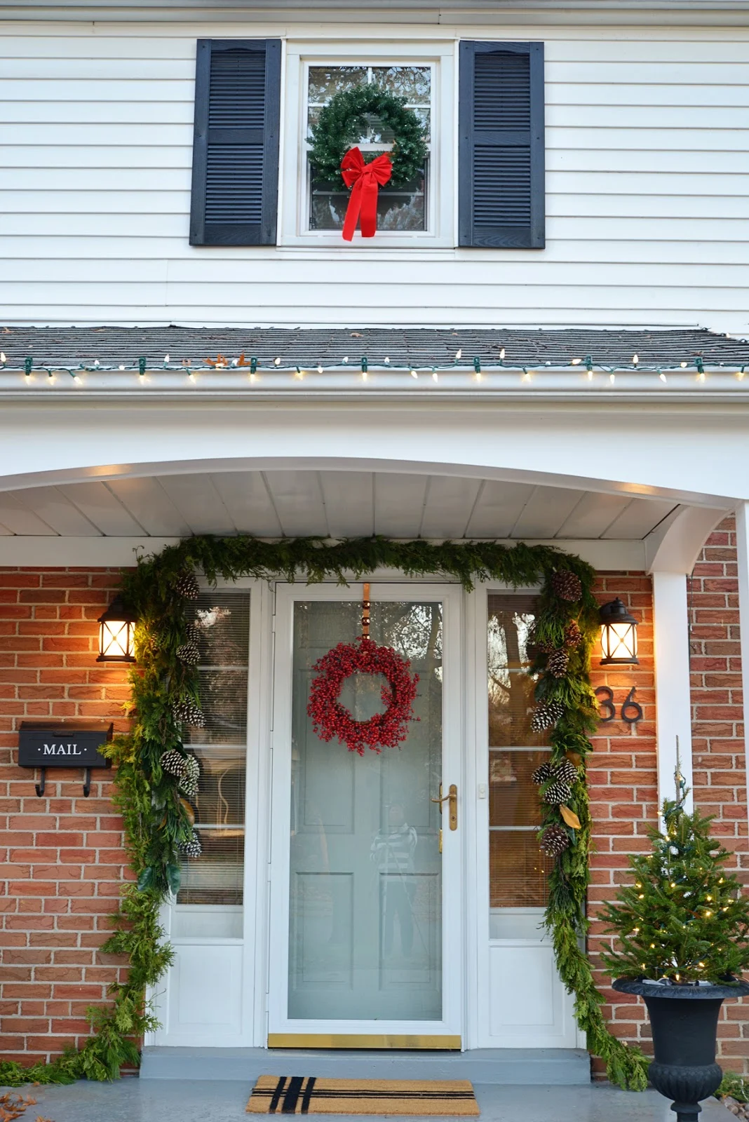red brick classic house, christmas garland around door, coastal outdoor lighting, mailbox with decal, trees in urn