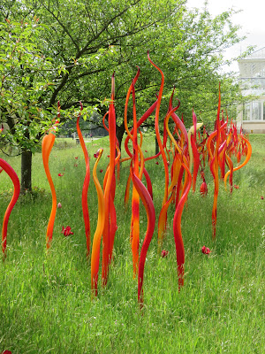 Photo of Chihuly glass sculpture - re and orange glass reeds standing in long grass under trees
