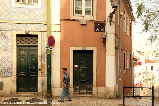 An elderly man in a checked shirt and a cap, walking past a row of colourful facades in the warm afternoon light.