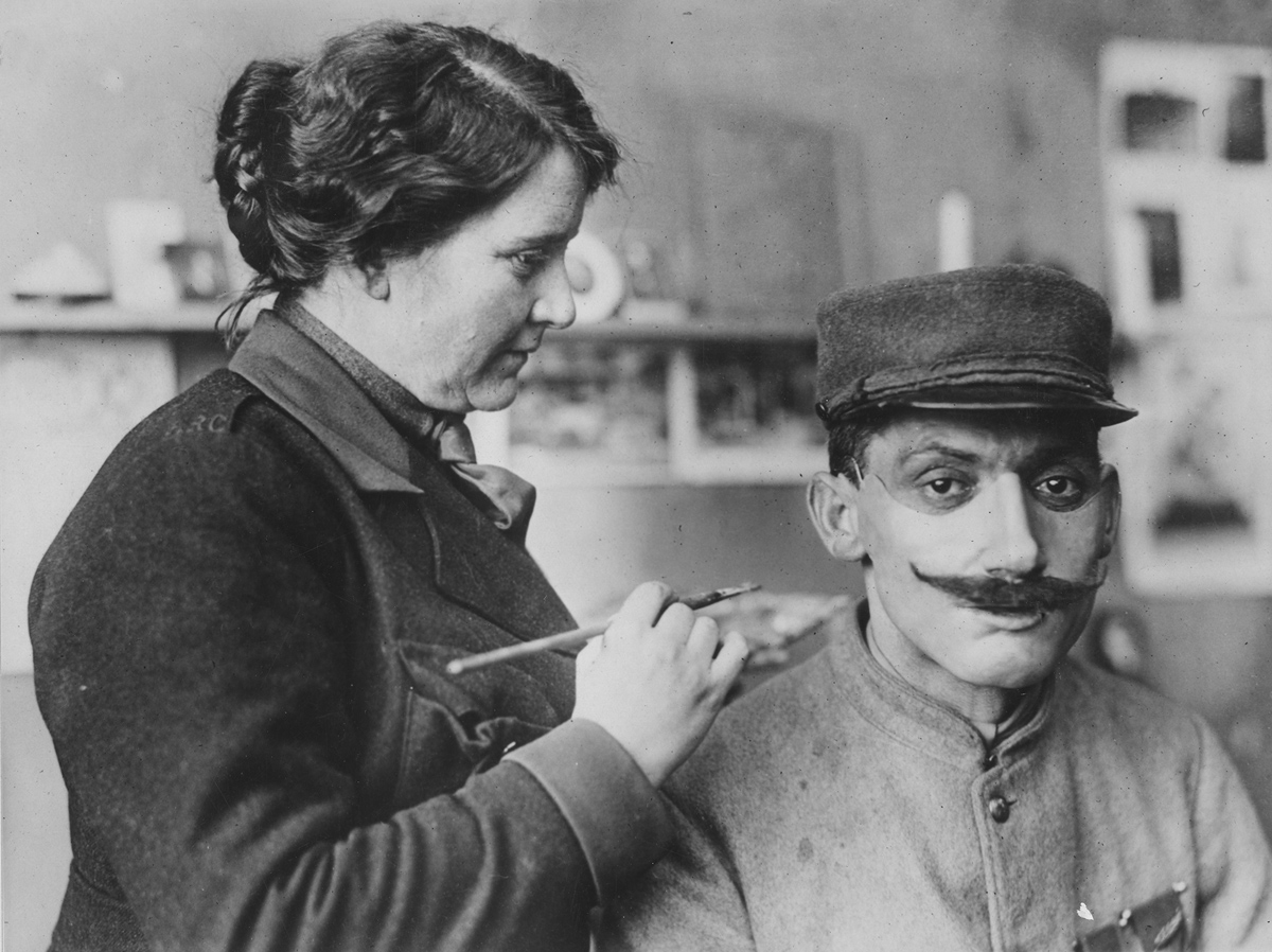 Masks made for men with disfigured faces from war wounds. Original caption: Mrs. Ladd coloring one of the masks after adjusting on a wounded Poilu's face.