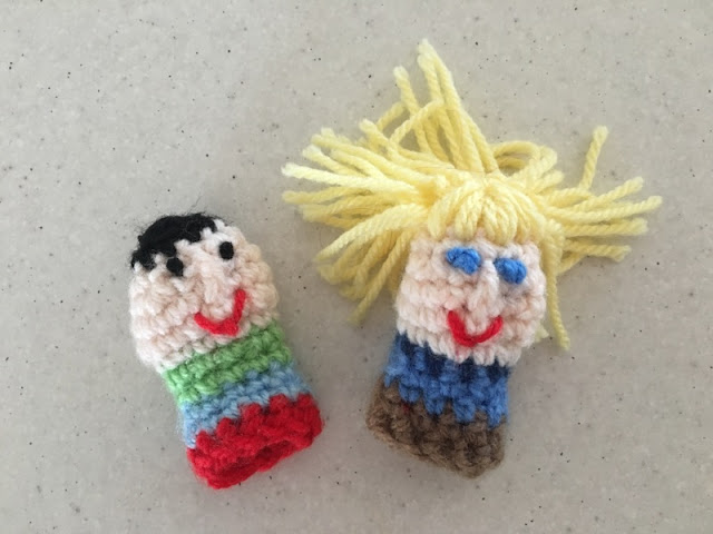 Two little finger puppets side by side on the benchtop. The left one has short black hair, a pale face with two black dotty eyes, a post stitch nose and a red embroidered mouth. The body is a striped crocheted cylinder striped from top to bottom: apple green, sky blue, red.  The right hand puppet has long yellow hair with satin stitch fringe, a pale face, blue oblong eyes, a post stitch nose and red embroidered mouth, The body is striped with a dark blue collar, then light blue and brown.