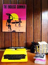 One-twelfth scale modern miniature office with wood-panelled wall, and wooden desk. On the desk is a yellow typewriter, set of yellow books and various pens and pencils in a glass.