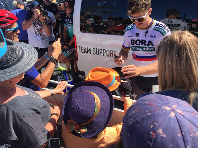 Peter Sagan stands in front of the tour bus to sign autoographs. I am holding out my orange souvenir hat and wearing a dark blue hat with an orange-tan and green silk ribbon.A crowd is in the foreground, a professional photographer can be seen on the top left hand quadrant of the picture behind the tour bus. Daniel Oss is wearing a black and green guernsey sitting on the bus tailgate.