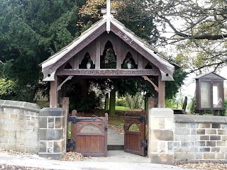 A wooden double gate set in a stone wall with an gabled structure above.  Decoratively carved and with shaped fretwork.  Surmounted by a wooden cross.