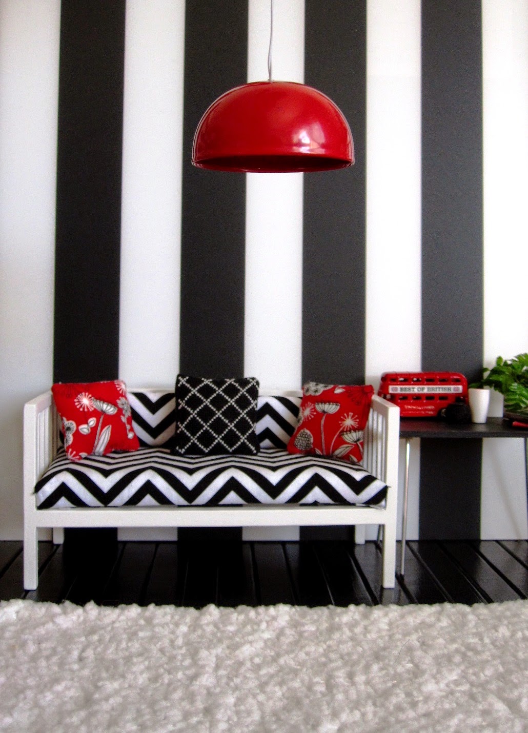 Modern miniature black white and red scene of a day bed with cushions next to a table displaying a model London bus, vases, books and a plant. A large red lightshade hangs from above and a white flokati rug is on the floor.