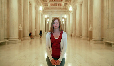 Photo of a 20-something white woman with sandy blond hair, standing in a big marble hallway