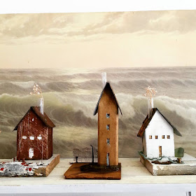 Three small assemblage-art houses on display in front of a picture of a stormy sea.