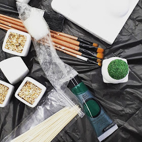 Flat lay of green paint, brushes, miniature platers and skewers against a black plastic background.