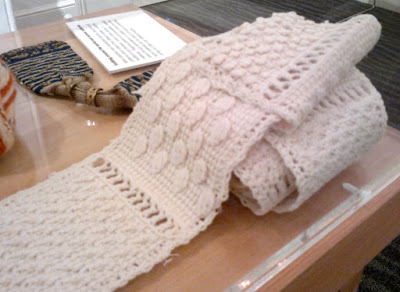 An off-white sampler scarf rolled up on the right hand side and laid out on the left. It is made up of squares separared by a row of filet openwork. Each square has a textured pattern worked in tricot (Tunisian crochet).