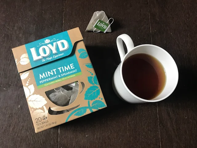 A white mug with mint tea in, a triangular tea bag and a box of Loyd The Magic Experience Peppermint and Spearmint tea