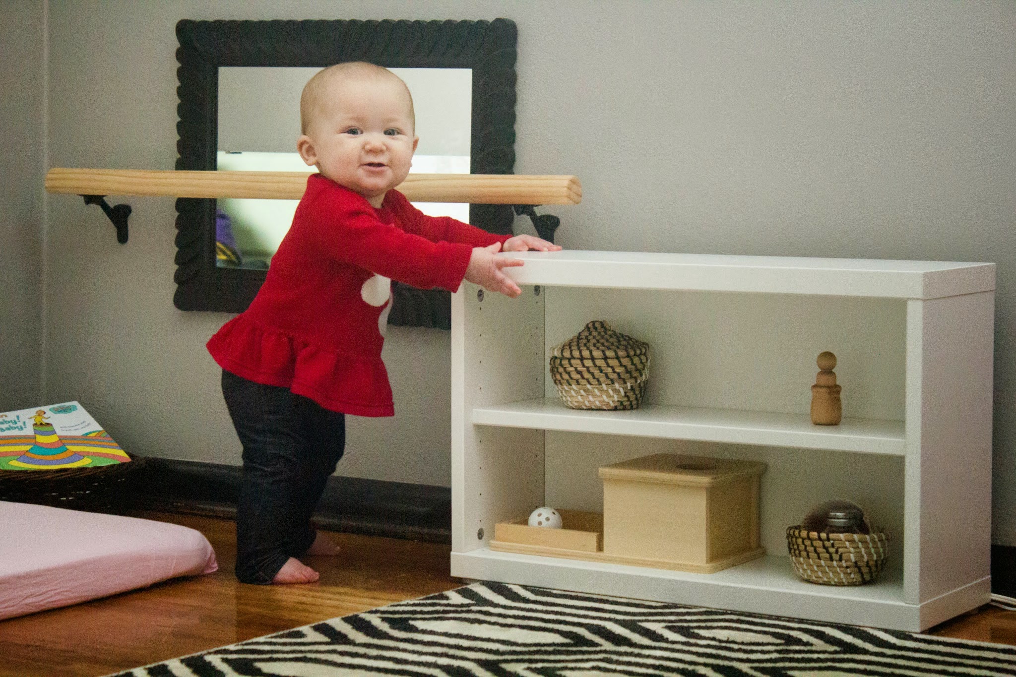 Montessori baby moves from pull-up bar to standing at shelf, she smiles to the camera as she is proud of her new movements.