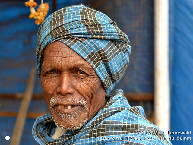 Closeup street portrait (outdoor headshot, seven-eighths view) of an old Indian Adivasi man, wearing a blue-checkered turban; Sanjay Market, Jagdalpur, Bastar District, Chhattisgarh, India. Matt Hahnewald Photography; Facing the World; photography; photo; image; face perception; physiognomy; psychological; Nikon D3100; Nikkor AF-S 50mm f/1.8G; 50mm lens; 4x3 aspect ratio; horizontal format; street; portraiture; closeup; headshot; seven-eighths view; blue; outdoor; colour; cultural; character; personality; real people; human head; human face; human eyes; bloodshot eyes; teeth; facial expression; lived-in face; wrinkles; eye contact; unshaved; stubbly beard; turban; headwrap; consent; empathy; rapport; encounter; respect; dignity; ethnic portrait; tourism; travel; travel portrait; market; Sanjay Market; Jagdalpur; Bastar District; Chhattisgarh; India; old man; Indian man; Adivasi man; one person; male; adult; senior; posing; authentic