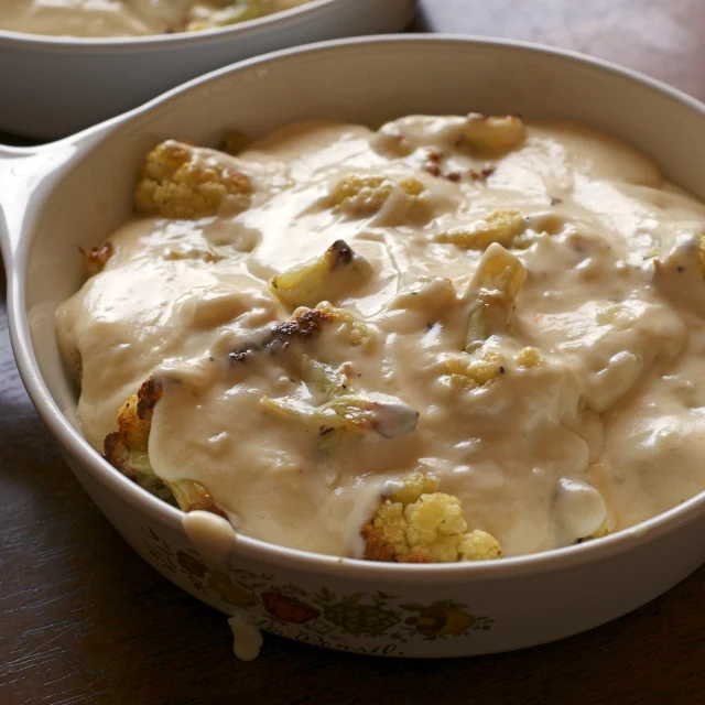Roasted cauliflower topped with melted white cheese sauce in a vintage white casserole dish on a wooden background.