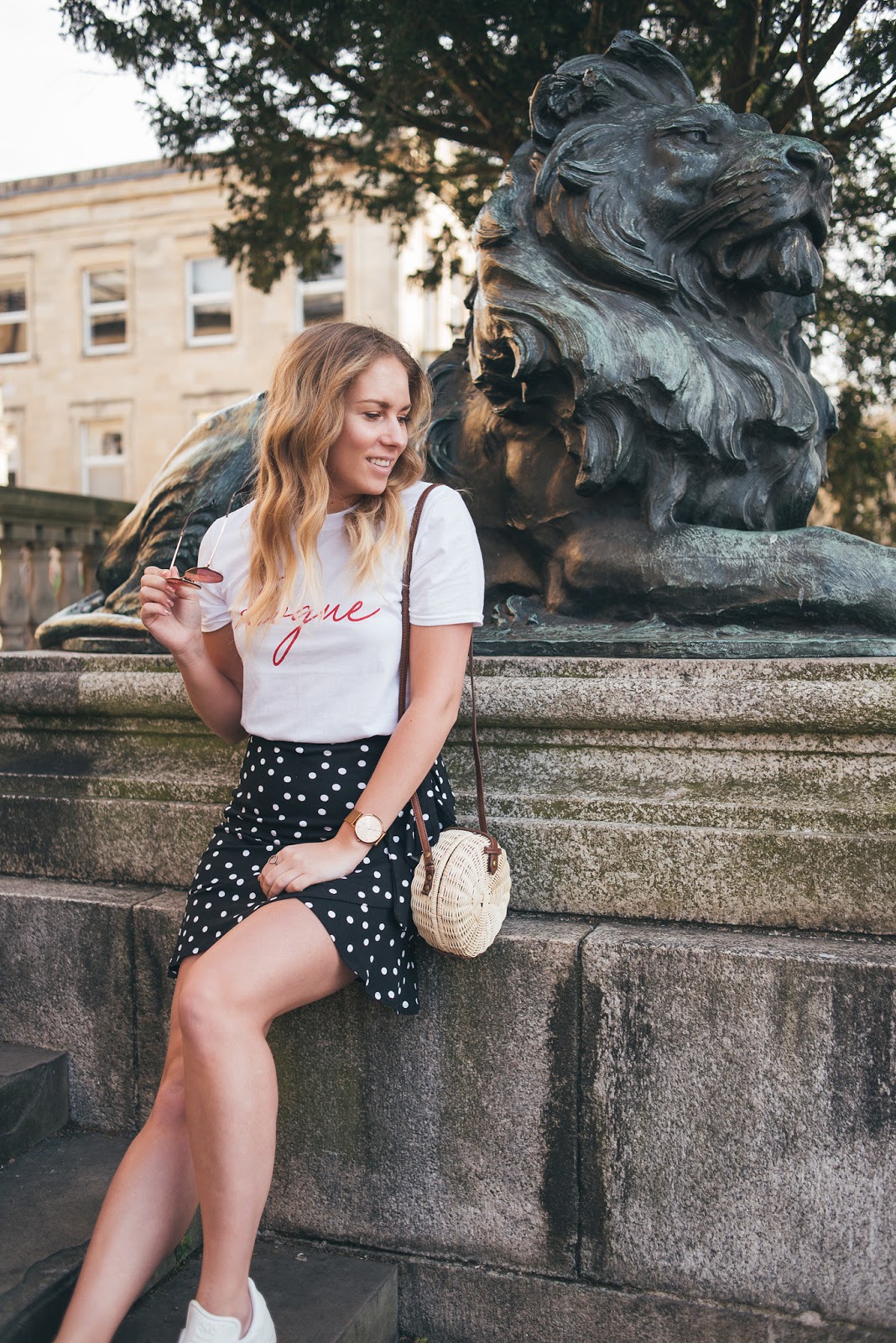 Close up of Rachel Emily sitting by Lion Statue in White Rogue Tee, ASOS Polka Dot Skirt with straw bag and ray ban pink blaze sunglasses