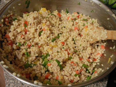 Couscous, chopped tomatoes, chopped bell peppers, chopped fresh cilantro, chopped green onion, in a metal bowl with a wooden spoon 