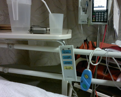 The view from my hospital bed clockwise from top left: overway table upon which is a water jug, transistor radio and glass of water with straw; infusion pump; over the sides of the bed hang the tubing for the drip, the nurse call bell and the bed controller.