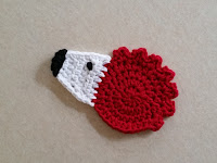 A 'hedgehog' motif consisting of a red circle crocheted in the round for the body with a jagged border across the top for spines; a white triangular face with black embroidered eye and nose.