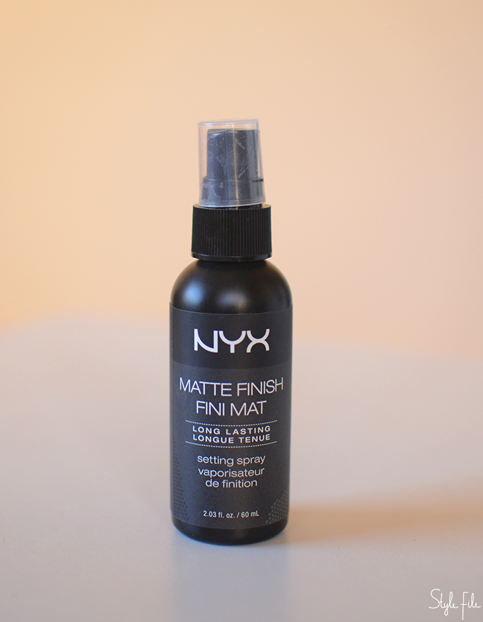 An image of NYX matte finish setting spray in a black spray bottle in front of a peach background for a beauty review