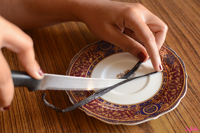 An image of a woman's hands holding a knife and slicing open a vanilla bean placed in a decorative plate on a wooden table to make a do it yourself project of how to make vanilla extract at home