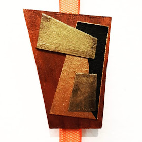 A brooch with geometric shapes in shades of copper, gold, bronze and black displayed on an orange netting ribbon.