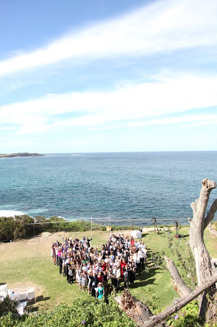 The wedding party on the cliff standing in a heart-shaped formation. One can see the view of the north head of Dee Why beach and the Pacific Ocean horizon in the distance.