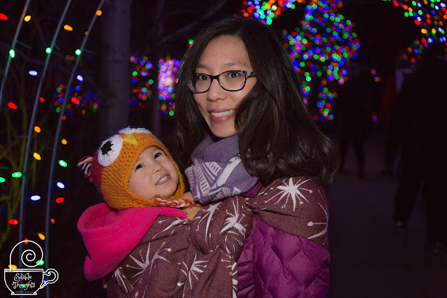 [Image of a smiling tan skin bespectacled Asian woman with dark brown hair wearing a toddler on her hip in a maroon and ecru star burst patterned woven wrap carrier over a purple down jacket. She also has on a purple and ecru windmill patterned infinity scarf made from wrap scrap. The toddler is smiling up at twinkling lights above. Toddler has on a pink fleece and a crocheted owl hat. They’re outside at night surrounded by colorful light decorations.]