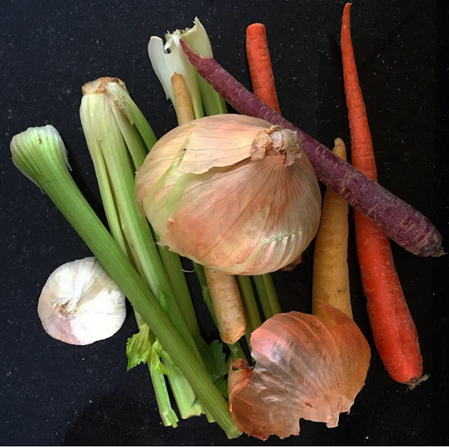 [Image of a white head of garlic, stalks of green celery, a yellow onion with peel artfully partially fallen off, an assortment of yellow/orange/purple carrots on a black kitchen counter.]