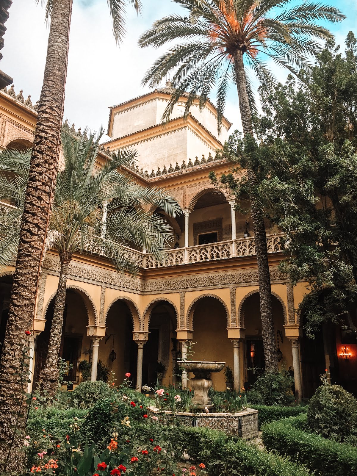 vue sur l'une des façades du palacio de las duenas à séville depuis le jardin avec au premier plan des arbustes en fleurs et une fontaine et à l'arrière plan la façade jaune du palais et ses arcades