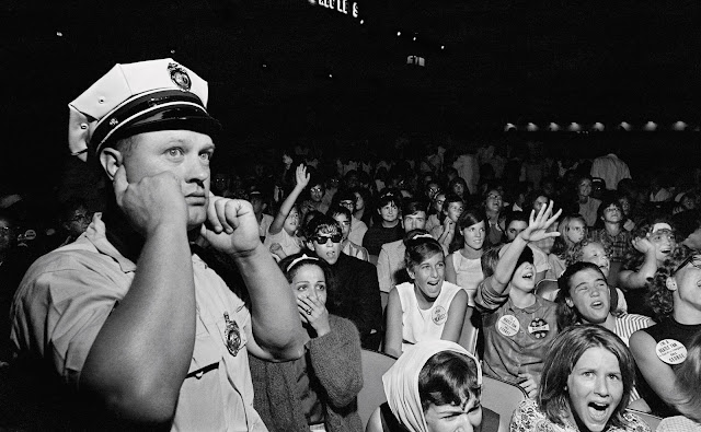 The Beatles, 1964 American Tour. Police officer with his fingers in his years with an audience of young girls screaming at the concert. Atlantic City, New Jersey Convention Hall. Pirate Radio and Sealand. Marchmatron.com The Beatles, 1964 American Tour. Police officer with his fingers in his years with an audience of young girls screaming at the concert. Atlantic City, New Jersey Convention Hall. Pirate Radio and Sealand. Marchmatron.com