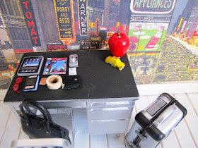 Modern dolls' house miniature desk with a selection of travel necessities arranged on it, including a tablet, guide books, digital camera, passport, glasses and a roll of duct tape.