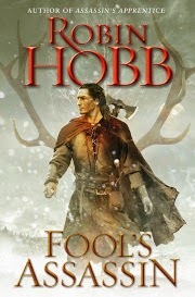 cover art for Fool's Assassin, featuring a pale-skinned man with long, dark hair walking through the snow. He has is head turned slightly to one side so he's in profile. He wears medievalesque leathers and carries an ax on his back. A large pair of antlers hover in the background.