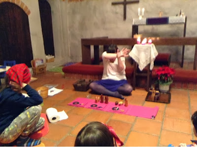 This is a modern-day photograph of a medieval chapel. A woman is seated on cross-legged on the floor, in front of the altar rail, facing away from the altar (towards the camera). She is wearing a white shirt, a purple skirt, and grey tights. She is white and has brown hair. There is a long narrow purple cloth on the floor in front of her, Among the objects laid on this cloth are some wooden figures from a Nativity Set (others are on a tray to her left). The woman has covered her eyes with her arms, as if to shield them from a bright light. Also in the photo are: a boy on the floor facing the Storyteller. He is wearing a Christmassy red stocking cap. We can just see the back of the head of a girl wearing a ribbon in her hair (facing the Storyteller) and the very edge of the face of a boy on the right also facing the Storyteller. In the left of the picture we also see, on the floor, a roll of paper towels, several baskets, and at least one piece of paper and some crayons.