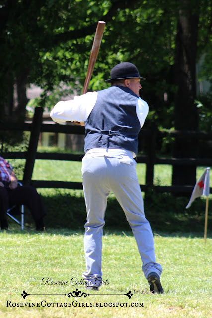 Man in profile. He is dressed in clothing from the 1800s and he has on a white dress shirt, dark blue vest and light blue pants with an old bowler hat on his head. He holds a baseball bat on one shoulder and is ready to swing the bat when the pitcher throws the ball. He is in a beautiful field with large trees.  The article is about vintage baseball by RosevineCottageGirls.com
