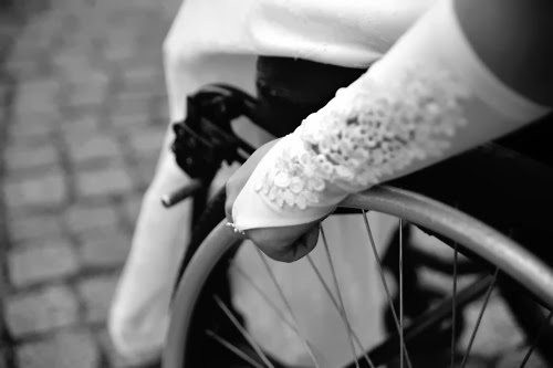 Black and white photo of a woman sitting in a manual wheelchair, closeup focus on her hand pushing the rim of the wheel, her hand wears a long white gloved with frills, looking like part of wedding dress