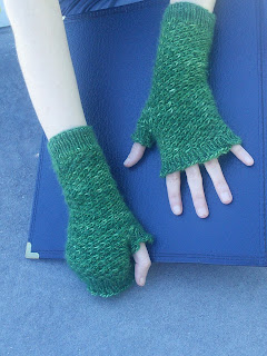 Someone wearing a pair of fingerless mittens. The mittens are knit in a dark green fingering-yarn and use a textured star stitch over the body of the mitten.