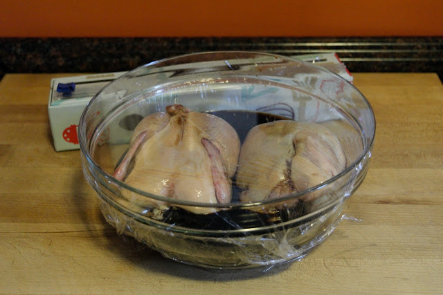 Two Cornish hens, in a bowl, with soy sauce, covered in plastic wrap. 