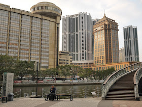 man using a mobile phone while sitting next to the Shiqi River with tall buildings in the background