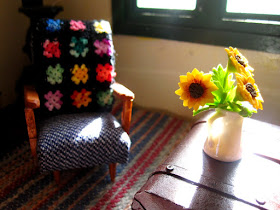 Modern one-twelfth scale miniature vintage armchair with crochet afghan over the back next to an old chest coffee table with a vase of sunflowers on it.