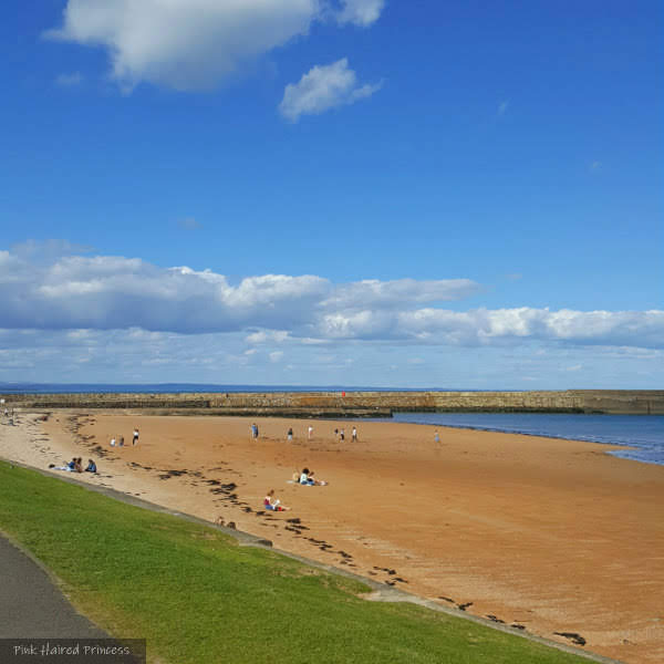 Summer FOMO with Myalgic Encephalomyelitis sandy beach, grass and pier in background with people sitting on Scottish beach in the sun