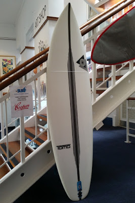 A surfboard leans against the staircase in the foyer of The Arts Centre. The surfboard is white with a black central stripe running from tip to tip. The brand logo Tomo is written on the left of the stripe and another logo is printed on the top right of the stripe.  The surfboard is standing vertically. It's cover which is solid grey with red piping edges is hung horizontally from the balustrade.  There is a small printed notice displayed to the left of the surfboard explaining the Tomo surfboard and cover together is the People's Choice prize donated by Coopers. The red Coopers logo is at the bottom and the Surf Art Exhibition logo is at the top of the notice. 