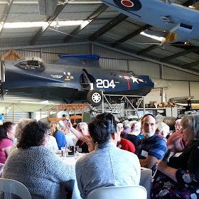 Group of people talking at a table in a hanger, with vintage airplanes suspended above.