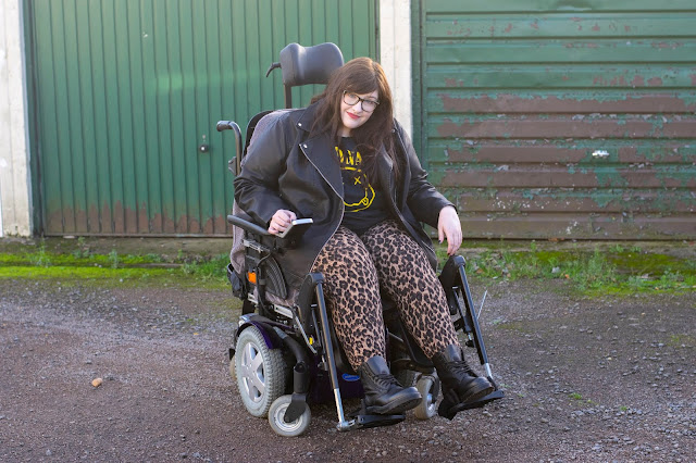 Sarah sat in powerchair with brown hair down wearing leopard print trousers, Dr Martens boots, leather jacket and Nirvana tee