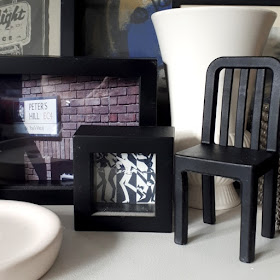 Miniature black chair on display amoungst a selection of framed pictures and white ceramic pieces.