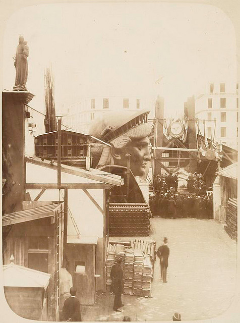 [View of the external area of the workshop in Paris, showing construction materials, the head of the Statue of Liberty, and a group of men gathered in front of the left foot of the statue.]. Fernique, Albert -- Photographer. 1883. Source: Album de la construction de la Statue de la Liberte. Repository: The New York Public Library. Photography Collection, Miriam and Ira D. Wallach Division of Art, Prints and Photographs.