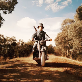 Model toy soldier riding a motorbike down a dirt road in an Australian bush setting.