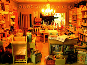 Ground floor of a modern miniature shabby chic shop, with a counter to the left, a dining table covered in candlesticks in the centre, wallpaper books to the back, fabric and paint on the right.