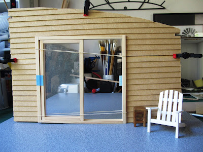 Wall of a dolls' house kit, with weatherboarding clamped to it,  and a sliding door dry fitted. The wall is standing on a piece of board that looks like concrete, and a dolls' house miniature Adirondack chair is in front of it.