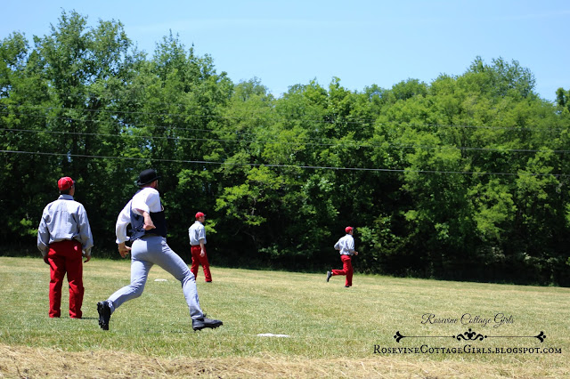 Men in a grassy field surrounded by large trees.  They are playing baseball and are wearing vintage uniforms from the 1860s.  The man in the blue uniform is running and almost to the base. Two of the men in the red uniforms are running for the ball.  The man on that base is watching the ball and waiting for them to throw it to him. The article is vintage baseball by rosevinecottagegirls.com
