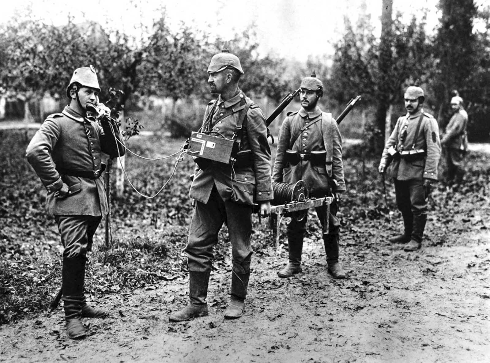 A German soldier holds the handset of a field telephone to his head, as two others hold a spool of wire, presumably unspooling it as they head into the field.